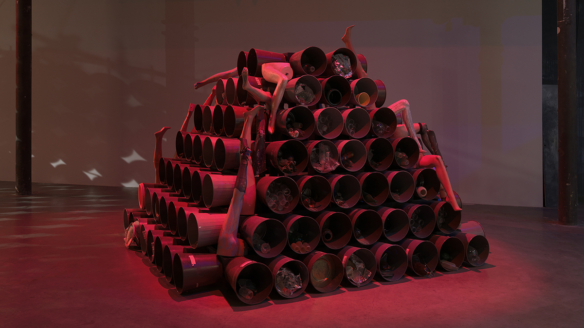 Color photograph of a pyramid shape sculpture comprising round plastic buckets and manniquin limbs underneath a ruby red cast of light