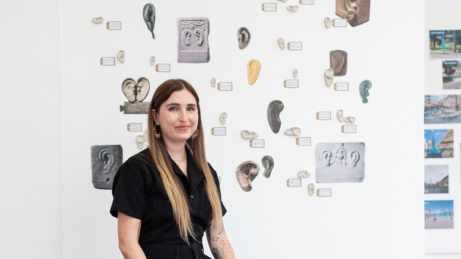 Color photograph of a light skinned woman with long straight hair wearing a dark jumpsuit; she stands in an artist's studio with tags and cutouts of ears affixed to the wall behind her