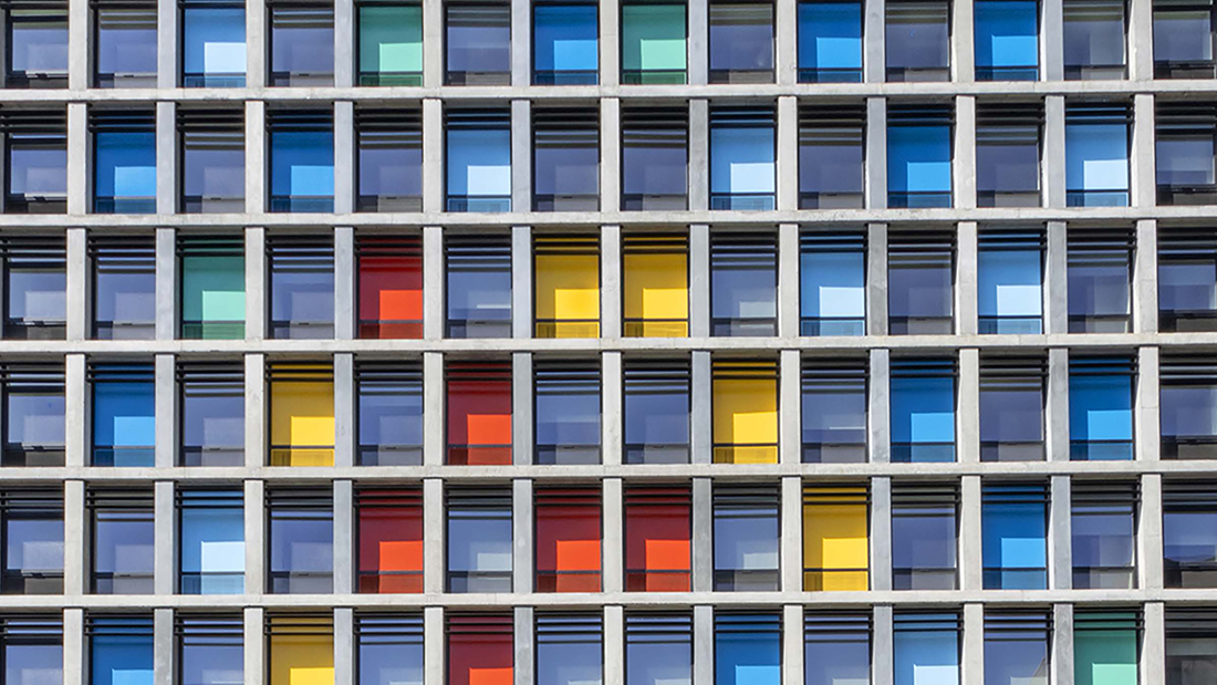 Color photograph of a detail of a modern building with gridded windows, with some windows colored light blue, red, and yellow
