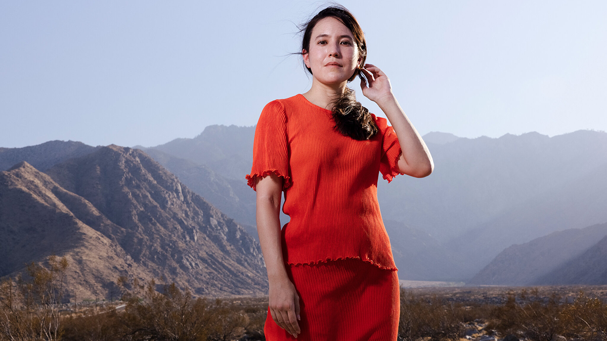 Color photograph of Alexandra Kleeman wearing orange clothes and standing in the Mojave Desert, with mountains behind her