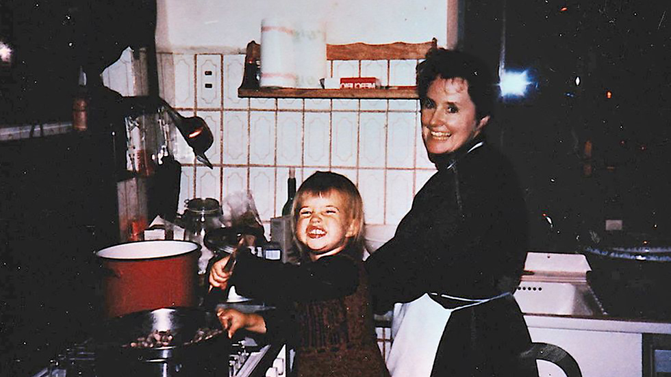 Vintage family color photograph of a woman and child in a kitchen cooking at the stove; both people look at the camera and smile