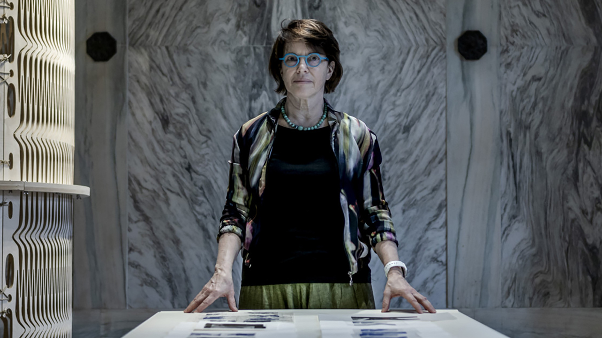 Color photograph of Anne Munly standing behind a table displaying two-dimensional work in her Syracuse exhibition