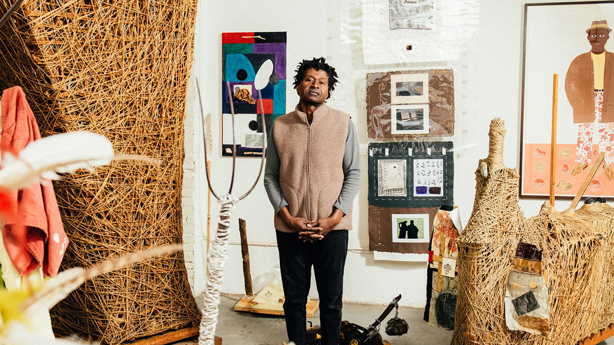 Color photograph of a dark-skinned man standing in an artist's studio surrounded by his sculpture, collages, and works on paper