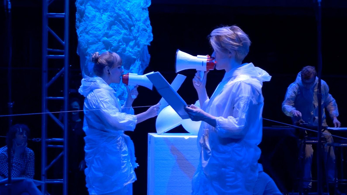 Color photograph of two woman onstage, each wearing a white gown and speaking through a megaphone while reading from a large sheet or notebook held in their hands; both women are bathed in blue light