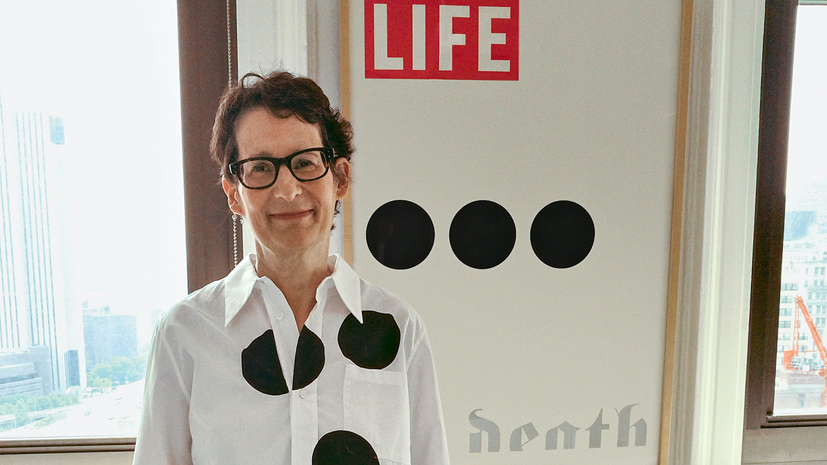 Color photograph of a light skinned woman wearing glasses and smiling at the camera; she is wearing a white shirt with three black dots, looking similar to an artwork behind her that has three similar black dots in a horizontal row