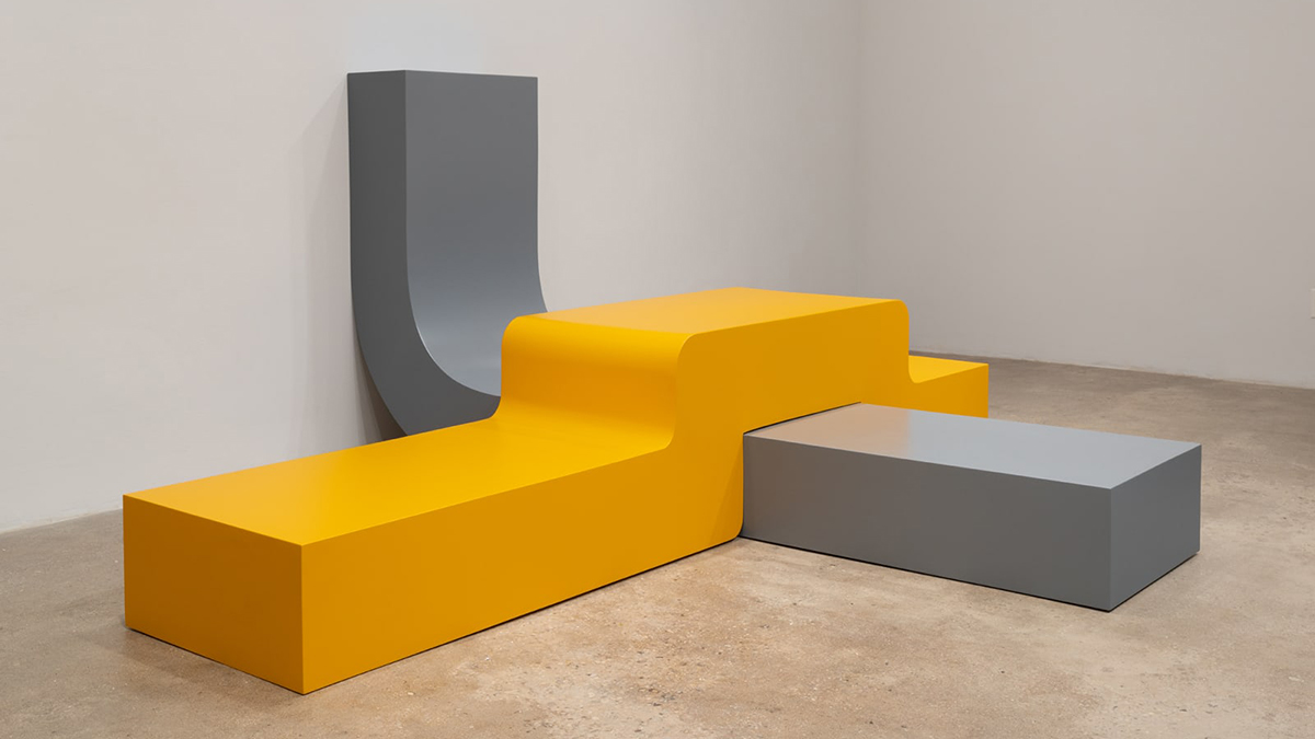 Color photograph of a gallery installation of curved, overlapping minimal sculpture, made of aluminum and painted yellow and gray, on the floor and leaning on the wall