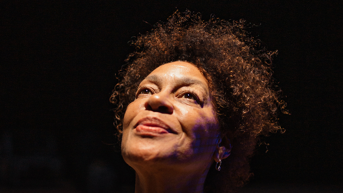 Color photograph of the head of a dark-skinned, brown-haired woman isolated against a solid black background; her face and gaze is tilted upward, spotlight from above