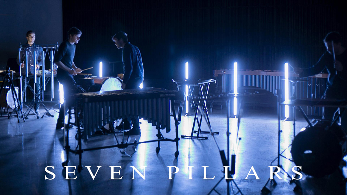 Color photograph of four men on a dark, indigo hued stage playing various percussion instruments; depicted are drums, marimbas, xylophones, and more