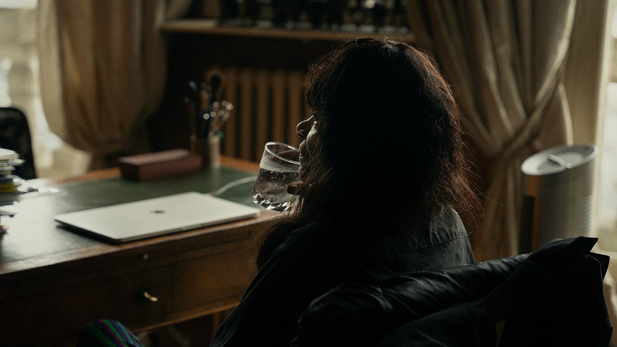 Color photograph of a dark skinned dark haired woman seen from behind at a 3/4 angle; she sits in front of a desk with a closed laptop on its surface