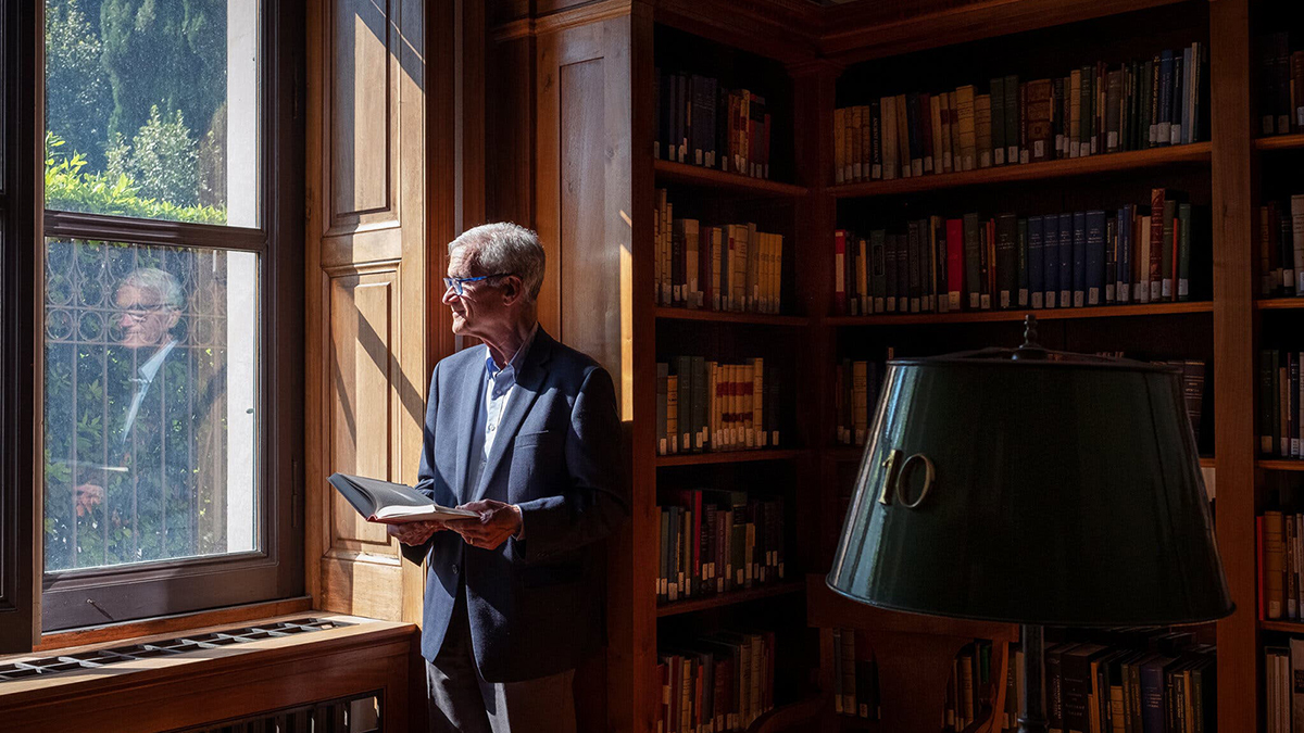Color photograph of an older light skinned man standing in a library with a book in his hand and books on wooden shelves behind him; he gazes out the large window