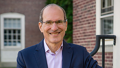 Color photograph of a light skinned man wearing a glasses, a blue blazer, and a light purple shirt, standing outside next to brick buildings, smiling and looking directly in the camera