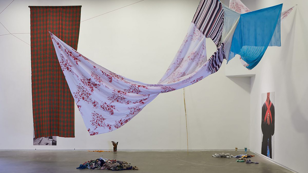 Color photograph of an artwork, consisting of colored, striped, and checkered fabric hung from the ceiling and across the room, installed in a white cube gallery