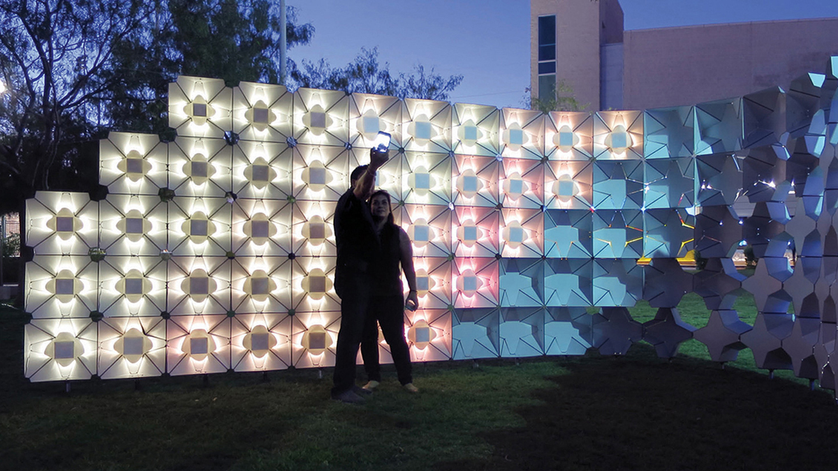 Color photograph of a man and woman taking a selfie at nighttime, standing in front of a temporary white-lighted latticed structure designed by Ersela Kripa and Stephen Mueller
