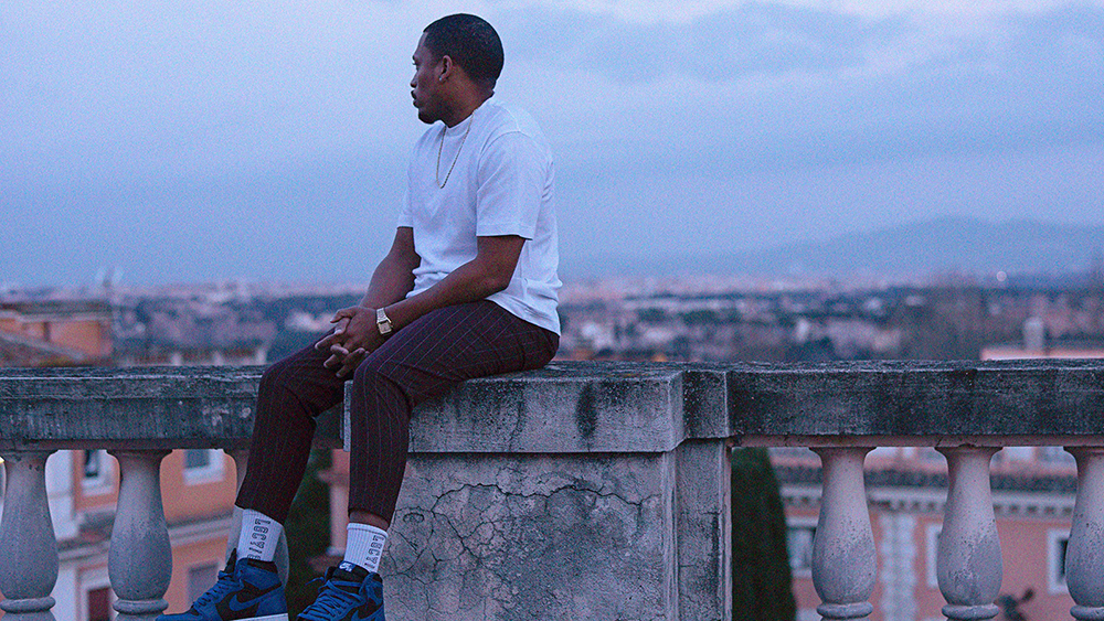Color photograph of a dark skinned man sitting in profile on a balcony ledge, looking out onto the Roman skyline at dusk