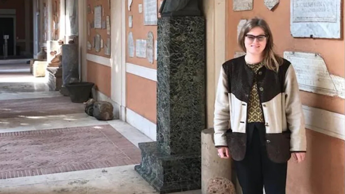 A light skinned woman standing in an arcade in the interior courtyard of the main American Academy in Rome building