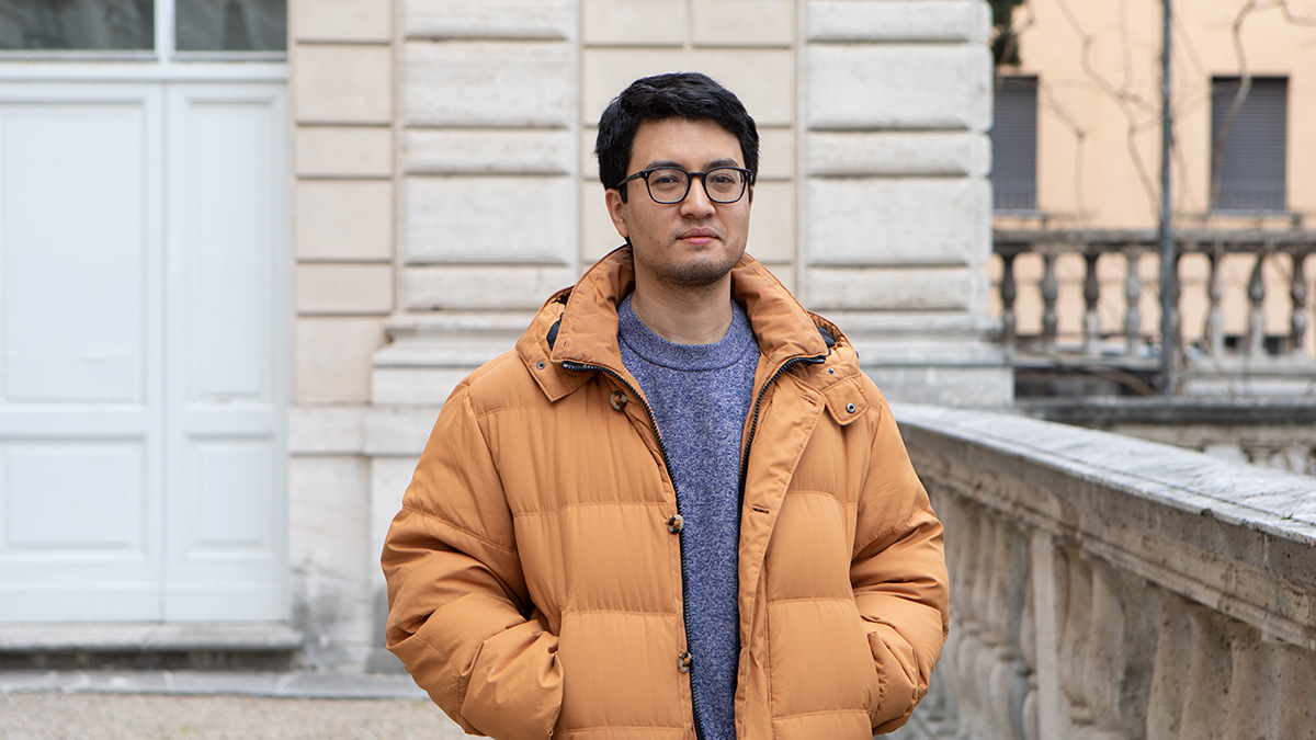 Color photograph of an Asian man wearing glasses and an orange puffer coat looking at the camera