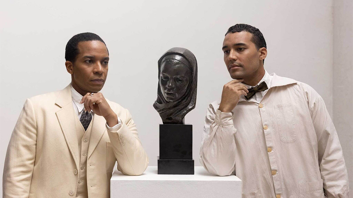 Color film still of two dark skinned men wearing white clothes and flanking a museum plinth that contains a sculptural bronze head of a woman