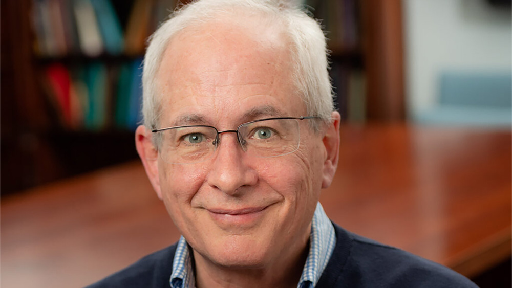 Color photo of the head and shoulders of an older light skinned man with white hair and glasses, smiling at the camera