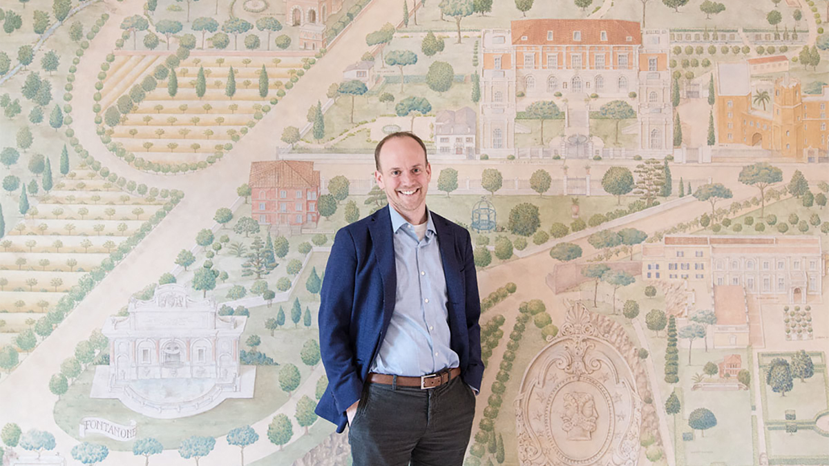 Color photograph of a light skinned man standing in front of a painted mural and looking at the camera