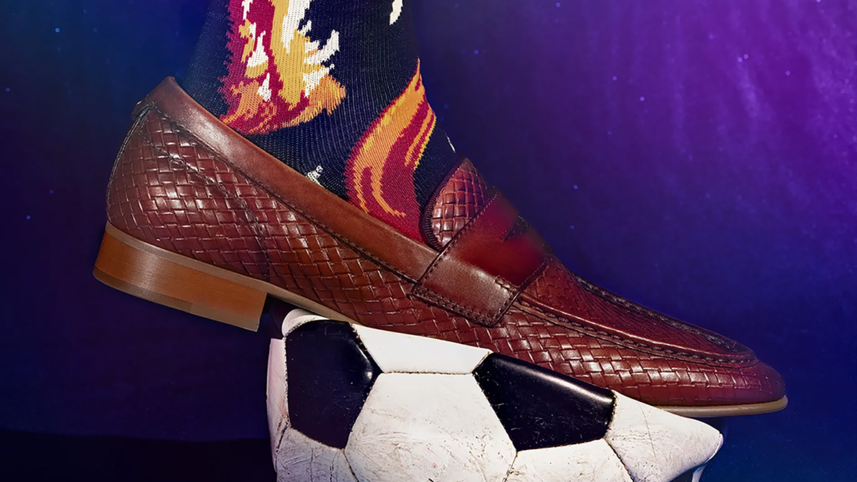 Color photograph of a human foot wearing a sock and shoe that are placed in top of a deflated soccer ball