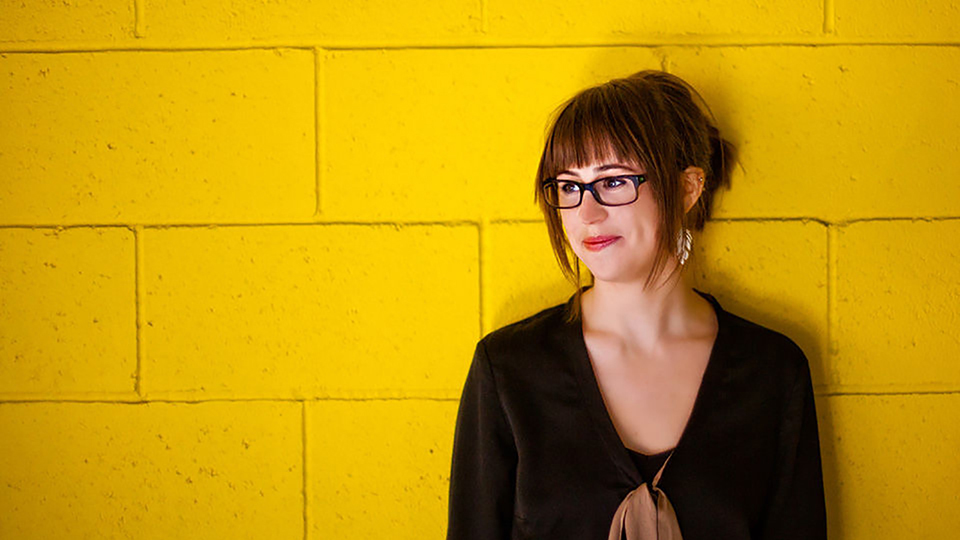 Color photograph of the head and torso of Katherine Balch standing against a cinderblock wall painted yellow