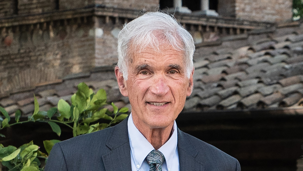 Color photo of the head and shoulders of an older white man wearing a suit and tie standing outside
