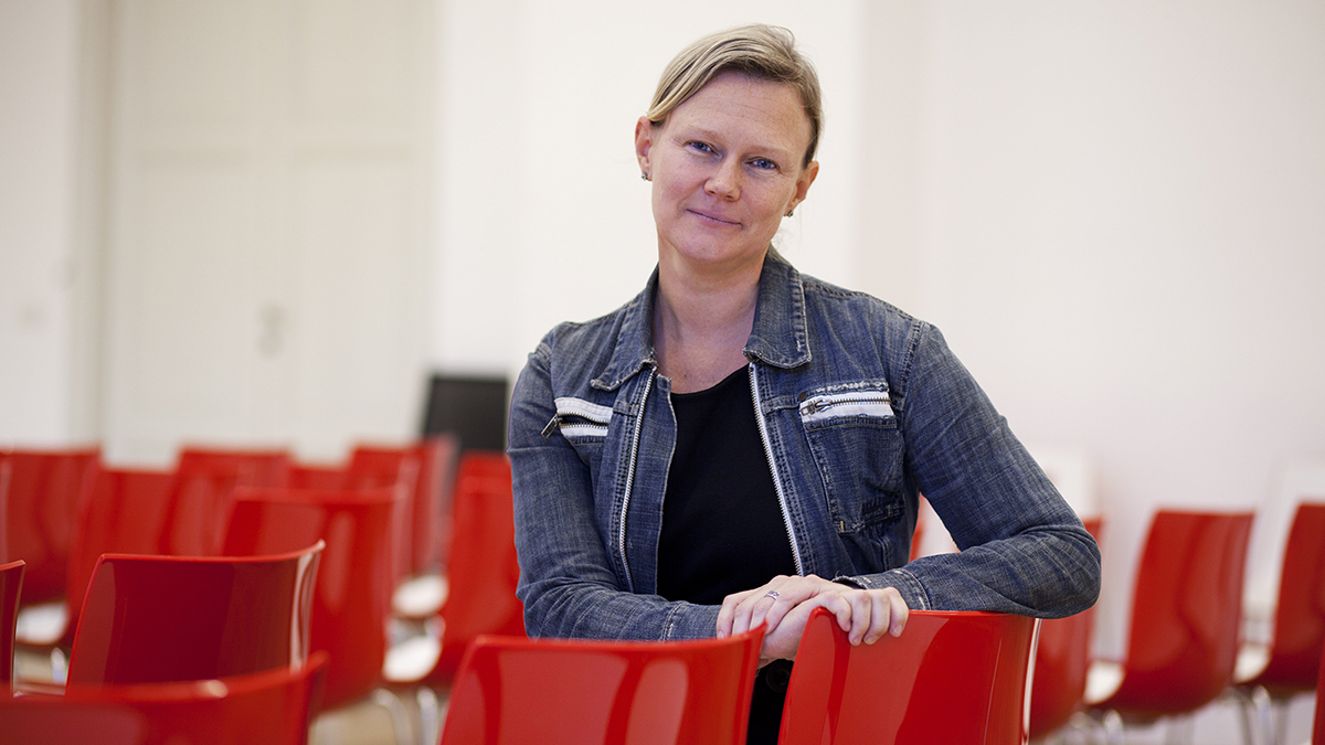 Color photograph of a light skinned woman with tied back blond hair looking at the camera with a slight smile; she sits in a red chair in a lecture hall