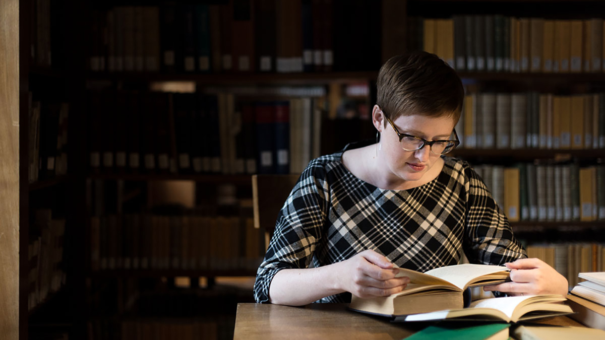 Color photograph of Lauren Ginsberg sitting at a table in the AAR Library, absorbed in reading a book