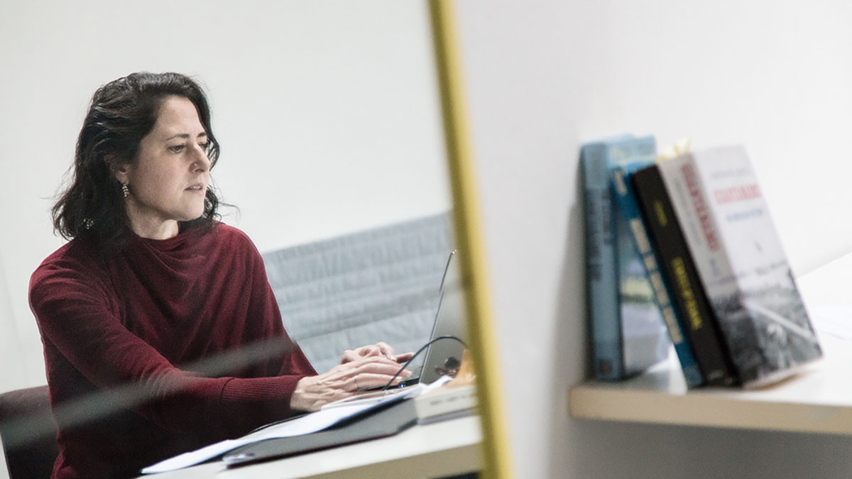 Color photograph of a light skinned woman sitting in front of a laptop as seen in a mirror; to the right of the mirror are three books on a shelf leaning against a wall