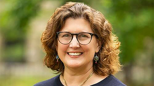 Color photograph of the head and shoulders of a light skinned woman with brown hair and wearing glasses smiling at the camera while standing in an outdoor garden like location