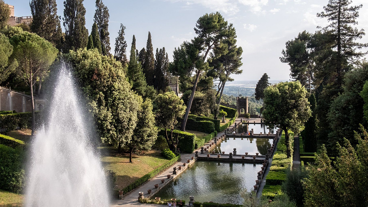 Color photograph of the Renaissance-era gardens at Villa d'Este, showing manicured trees and bushes flanking a long pool with a spouting fountain