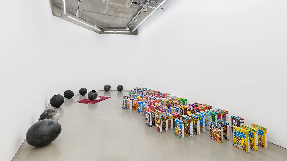 Color photograph of an art installation in a white-walled gallery; on the left eight black egg shaped sculptures; on the right are several dozen cereal boxes standing upright