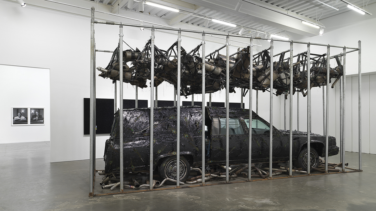 Color photograph of a white-walled museum installation of an actual hearse covered in black tar and peacock feathers; the automobile is situated in a cage made of steel poles, with mufflers in netting hanging above the car
