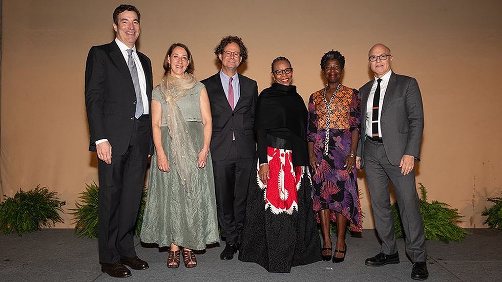 Color photograph of six men and woman wearing fancy suits and dresses standing in a row against a beige backdrop and potted ferns
