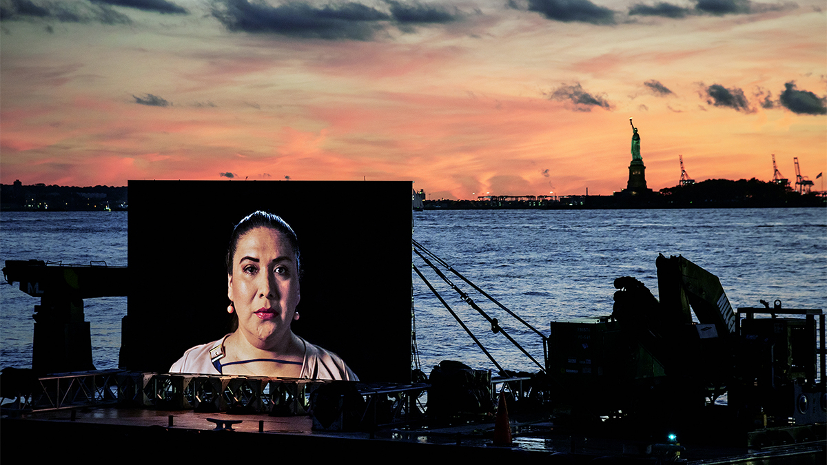 Color photograph of an artwork consisting of a large television screen (showing the head and shoulders of a woman) mounted on a barge in New York Harbor; behind the barge is the water, sky, and the Statue of Liberty