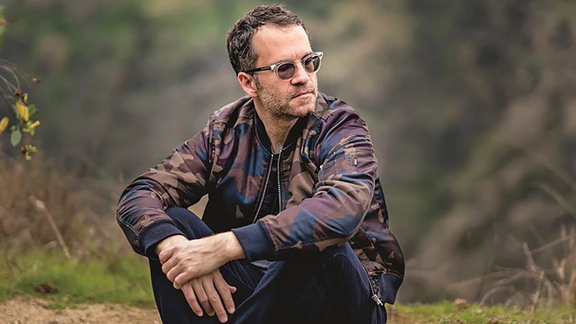 Color photograph of a light-skinned brown-haired man sitting cross legged in an outdoor location, looking to his left; he wears blue jeans, a camouflage-style jacket in brown, blue, and purple, and sunglasses