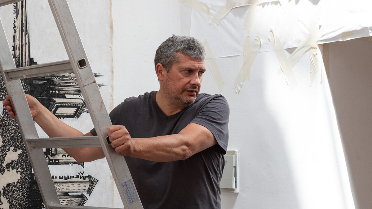 Color photograph of a light skinned man with graying hair and a gray t-shirt; he has descended a ladder in his art studio and looks off camera