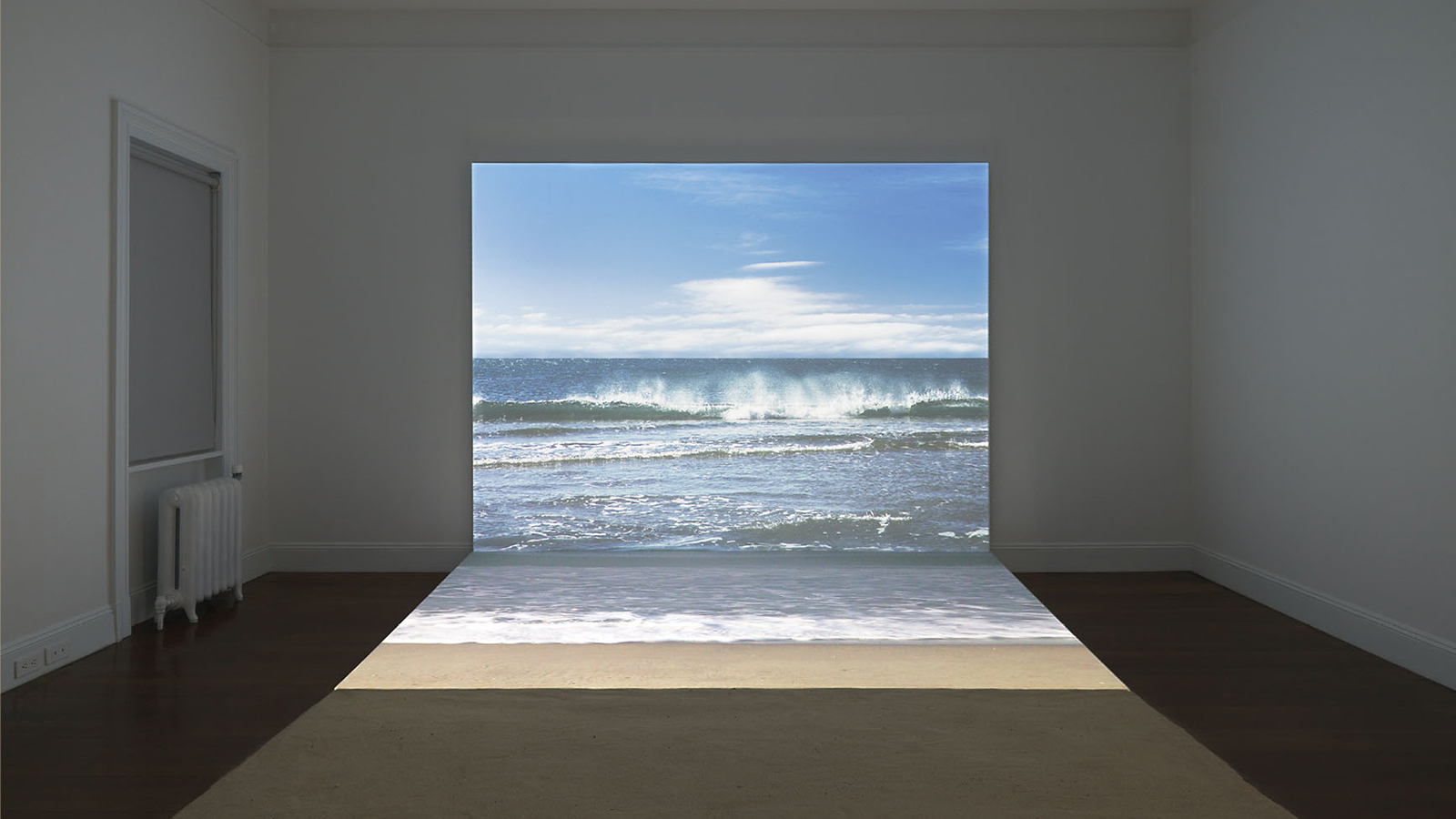 Detail of a book cover that has a color photograph showing the darkened interior of a gallery space with a video projected on the wall; visible on the screen are ocean waves crashing on a beach; on the gallery floor is a square of sand perpendicular to the image