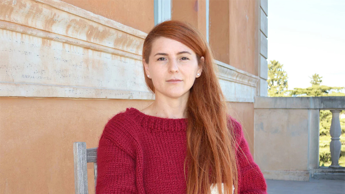 Color photograph of a light skinned woman with red hair wearing a red sweater, sitting on a rooftop patio in Rome
