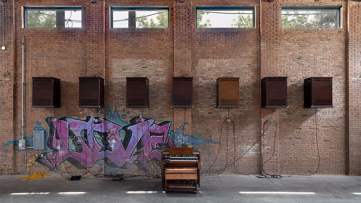 Color photo of an organ inside a warehouse in front of seven loudspeakers mounted on the wall