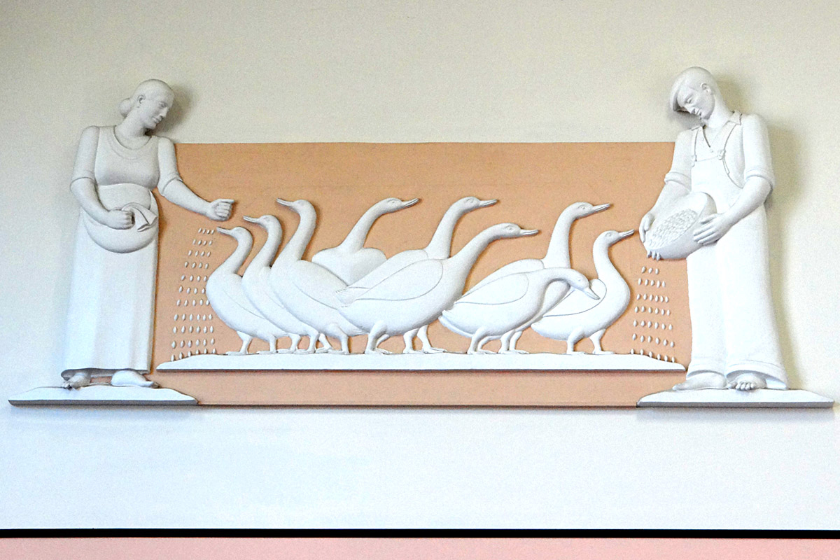 A wall mounted plaster cast sculpture shows two farmers, a woman on the left and a man on the right, feeding grain to nine geese between them