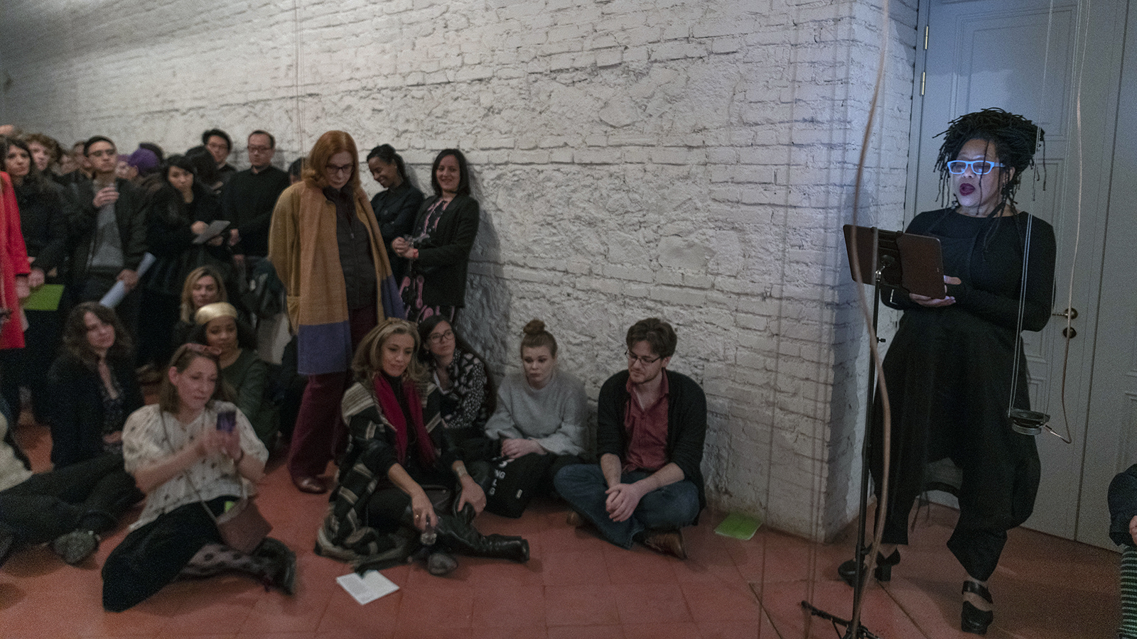 Color photograph of Pamela Z standing in front of a music stand and looking at a score on the left, and audience members sitting and standing to the right