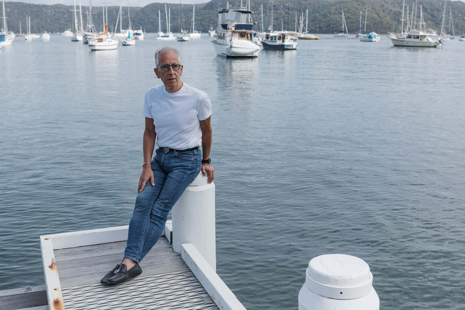 Color photograph of Simon Mordant wearing blue jeans, white t-shirt, and glasses and leaning on a dock pillar, with boats on a lake in the background