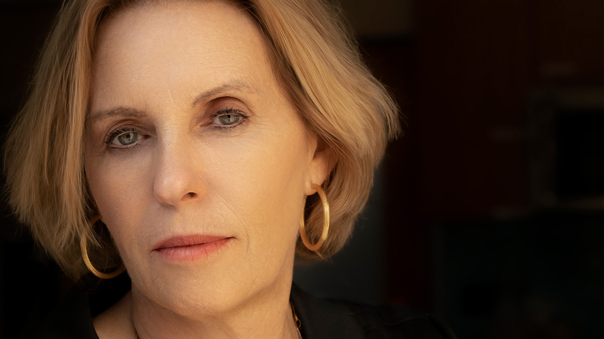 Color photo of the face of a light skinned woman with blond hair looking at the camera with a serious expression