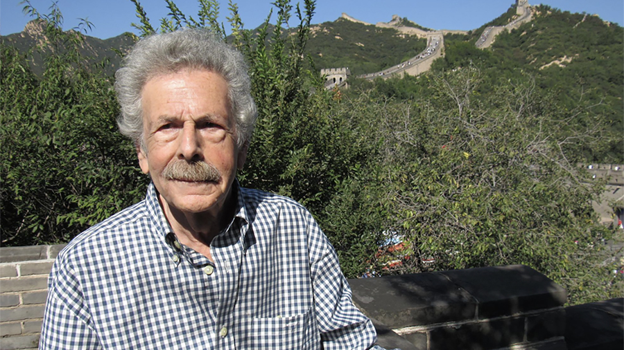Color photograph of an older, light skinned man looking at the camera as he stands in a green outdoor location