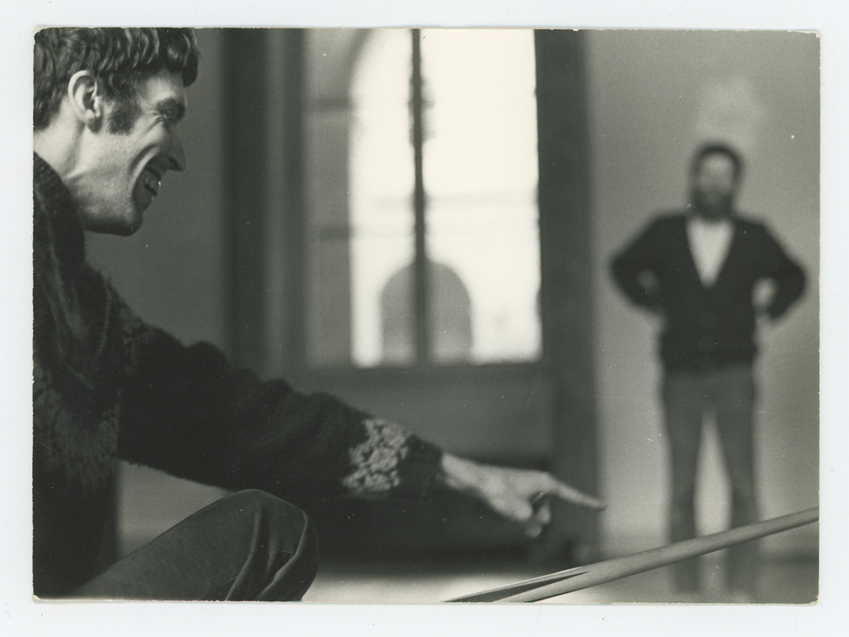 Black and white photo of a light skinned man kneeling and smiling; away from him and out of focus is a standing light skinned man