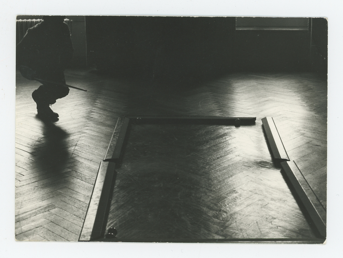 Black and white photo of a darkened room with wood slat floors; a person with a pool cue approaches a billiards &quot;table&quot; on the floor without a tabletop or legs