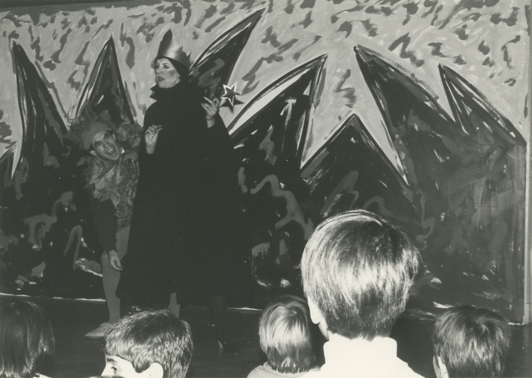 Black and white photo of two performers (man and woman) in costume standing in front of an abstract painted backdrop