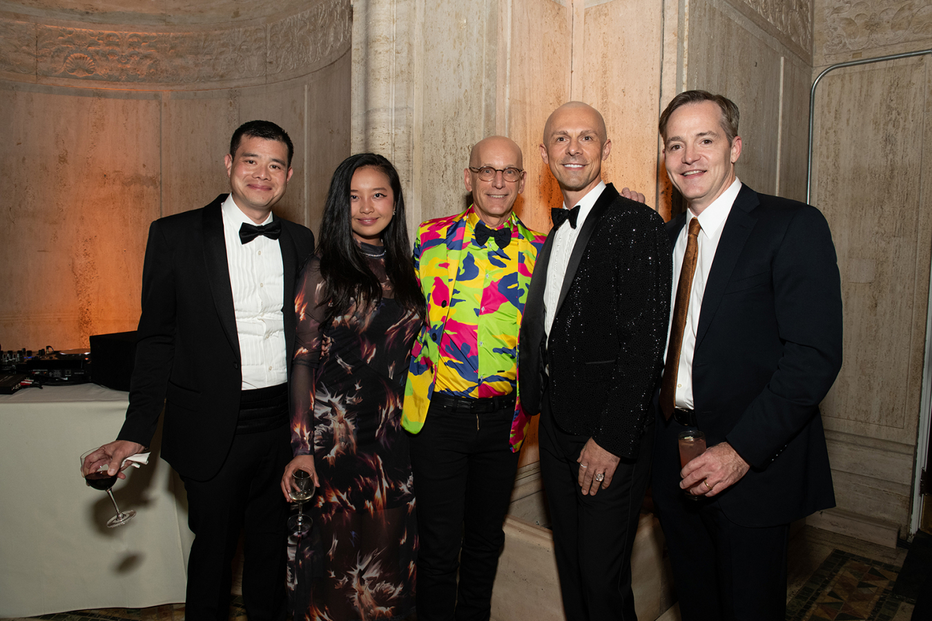 Color photograph of four men and one woman at a fundraising gala: there are two medium skinned and light skinned people standing in a row, wearing tuxedos, suits and ties, and a dress