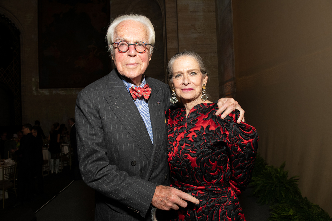 Color photograph of two light skinned elderly people at a fundraising gala: he wears a gray pin-striped suit and a bow tie; she wears a fancy black and red embroidered dress